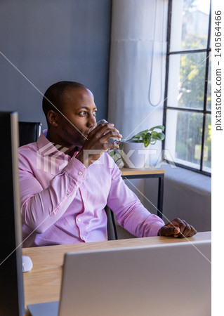 African american man in office sipping water from glass by laptop and monitor African american man in office sipping water from glass by laptop and monitor 140564466