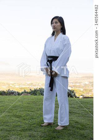 Asian woman standing barefoot on grassy hilltop practicing karate in white gi with belt Asian woman standing barefoot on grassy hilltop practicing karate in white gi with belt 140564482