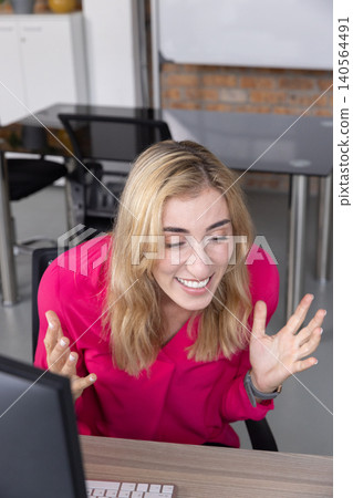 Woman in pink blouse leaning forward at desk in office raising hands with computer setup Woman in pink blouse leaning forward at desk in office raising hands with computer setup 140564491