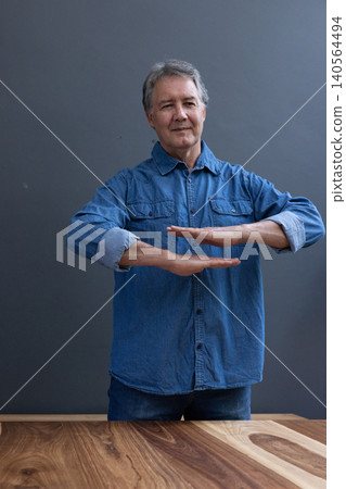 Senior man standing behind wooden tabletop holding hands parallel above grain patterns in room 140564494
