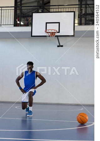 Kneeling african american male in blue jersey stretching on court near hoop with basketball 140564512