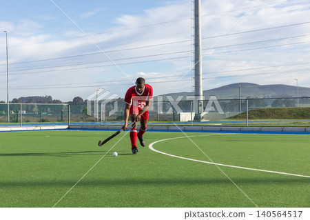 African american man in red uniform dribbling ball with hockey stick on turf at pitch African american man in red uniform dribbling ball with hockey stick on turf at pitch 140564517