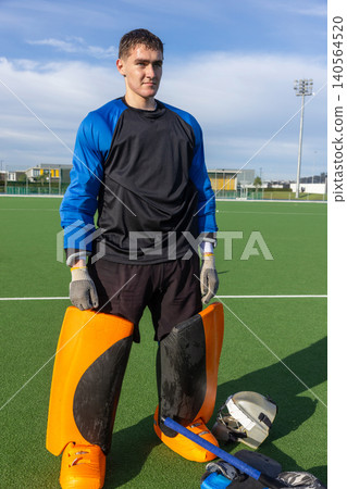 Male teenage goalkeeper standing on synthetic turf with padded gloves near helmet and stick Male teenage goalkeeper standing on synthetic turf with padded gloves near helmet and stick 140564520