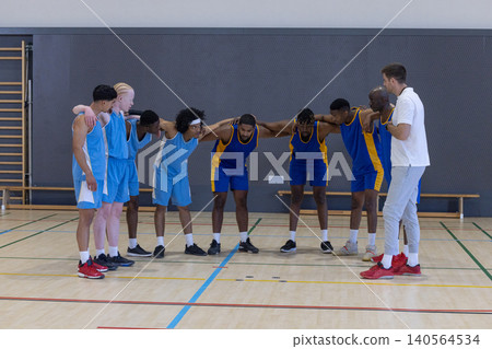Diverse male basketball team huddling around coach on court with jerseys Diverse male basketball team huddling around coach on court with jerseys 140564534