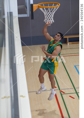 Man jumping under basketball hoop and net inside gym wearing green uniform with trim Man jumping under basketball hoop and net inside gym wearing green uniform with trim 140564536
