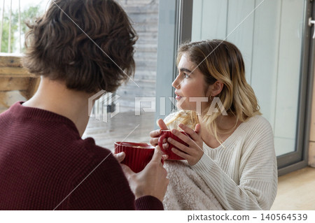 Couple wrapped in blanket sitting beside sliding glass door on floor holding red mugs and chatting Couple wrapped in blanket sitting beside sliding glass door on floor holding red mugs and chatting 140564539