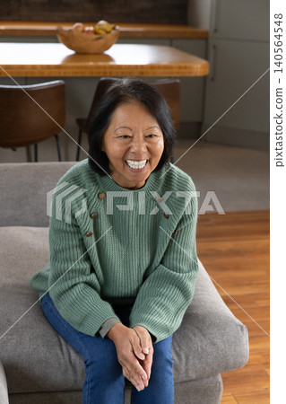 Senior Asian woman sitting on grey ottoman in living and dining area holding wooden fruit bowl Senior Asian woman sitting on grey ottoman in living and dining area holding wooden fruit bowl 140564548