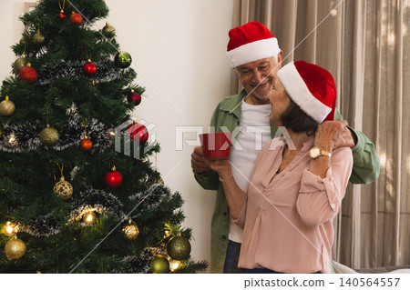 Senior couple wearing Santa hats standing at home by Christmas tree holding red mug, copy space Senior couple wearing Santa hats standing at home by Christmas tree holding red mug, copy space 140564557