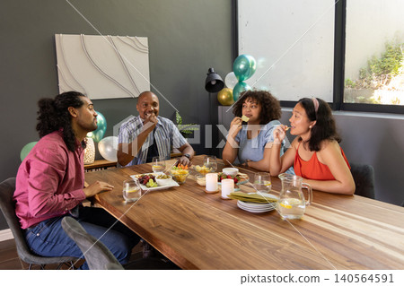 Diverse friends gathering in modern dining room around table sharing snacks under balloons Diverse friends gathering in modern dining room around table sharing snacks under balloons 140564591