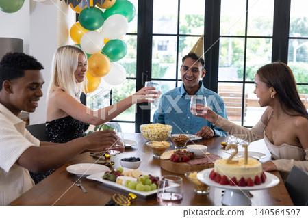 Diverse friends wearing party hats around wooden table celebrating birthday with cake, balloons 140564597