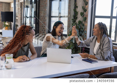 Diverse female coworkers celebrating milestone at conference table in office with silver laptop Diverse female coworkers celebrating milestone at conference table in office with silver laptop 140564598
