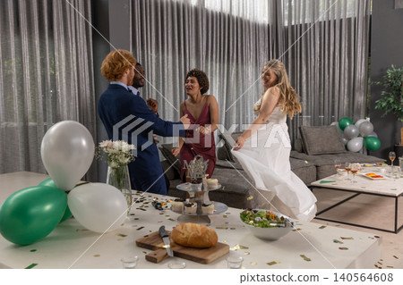 Diverse group of four in wedding attire dancing at home with confetti, balloons, champagne 140564608