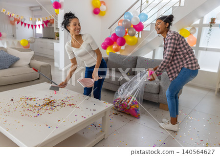 Diverse mom and teenage daughter sweeping confetti into dustpan while holding trash bag at home Diverse mom and teenage daughter sweeping confetti into dustpan while holding trash bag at home 140564627