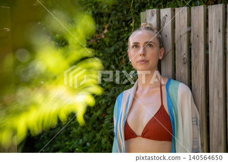 Female standing against wooden fence in garden wearing rust-colored bikini top, wrap, copy space Female standing against wooden fence in garden wearing rust-colored bikini top, wrap, copy space 140564650