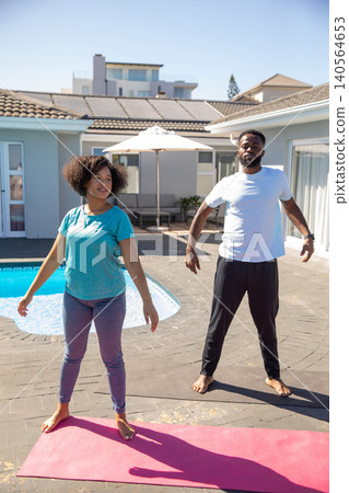 Diverse couple wearing sportswear practicing stretching exercises on yoga mats, backyard pool 140564653