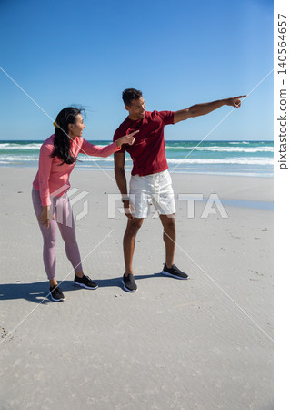 Diverse couple leaning forward and pointing toward ocean horizon on sandy beach with shoes Diverse couple leaning forward and pointing toward ocean horizon on sandy beach with shoes 140564657