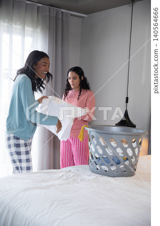 Diverse female friends folding laundry in bedroom using gray laundry basket, white duvet Diverse female friends folding laundry in bedroom using gray laundry basket, white duvet 140564666