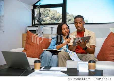 African American couple sitting on sofa in living room reviewing tablet with notebook and paperwork African American couple sitting on sofa in living room reviewing tablet with notebook and paperwork 140564695