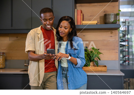 African American couple leaning against countertop in kitchen by potted plant looking at smartphone African American couple leaning against countertop in kitchen by potted plant looking at smartphone 140564697