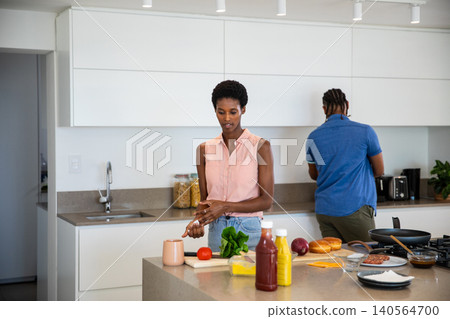 Diverse couple chopping tomato and greens while brewing coffee at kitchen island Diverse couple chopping tomato and greens while brewing coffee at kitchen island 140564700