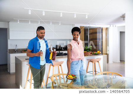 African American couple carrying plates of burgers and colored drinks to glass-top table in kitchen 140564705