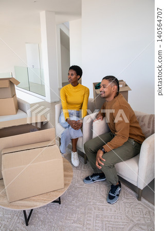 Diverse couple sitting on cream armchairs unpacking boxes on coffee table in modern living room Diverse couple sitting on cream armchairs unpacking boxes on coffee table in modern living room 140564707