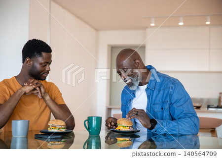 African american father and son sitting at glass dining table in kitchen with burgers, mugs African american father and son sitting at glass dining table in kitchen with burgers, mugs 140564709