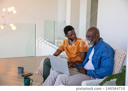 African american father and son sitting on sofa in living room using laptop, coffee mugs African american father and son sitting on sofa in living room using laptop, coffee mugs 140564710