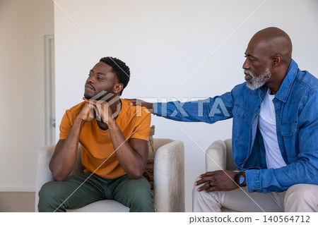 Senior african american father placing hand on adult son's shoulder on armchairs at home Senior african american father placing hand on adult son's shoulder on armchairs at home 140564712
