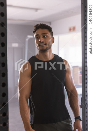 Adult indian male resting near steel rack in gym, wearing black tank top, smartwatch and shorts Adult indian male resting near steel rack in gym, wearing black tank top, smartwatch and shorts 140564803