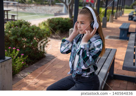 Female child adjusting wireless headphones on bench under pergola over brick pavers Female child adjusting wireless headphones on bench under pergola over brick pavers 140564836