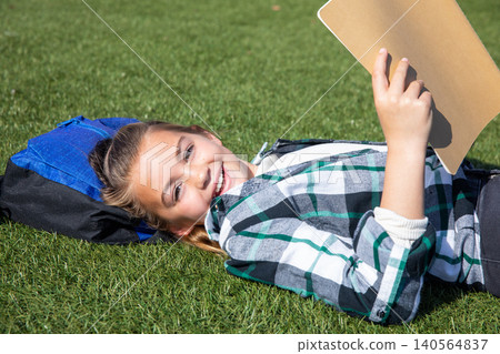 Female child lying on grassy field using blue and black backpack as pillow with tan clipboard Female child lying on grassy field using blue and black backpack as pillow with tan clipboard 140564837
