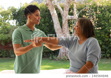Senior woman and male caregiver guiding arm stretch on white bench by trimmed hedge Senior woman and male caregiver guiding arm stretch on white bench by trimmed hedge 140564846
