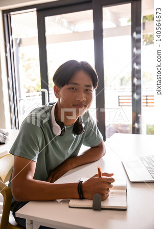 Asian male teenager writing in notebook with pen at desk near laptop and headphones Asian male teenager writing in notebook with pen at desk near laptop and headphones 140564873