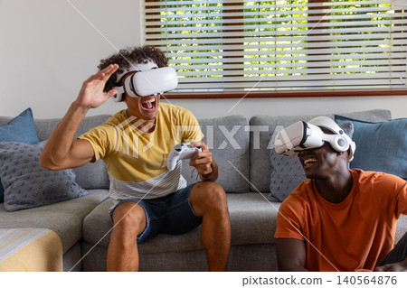 Diverse male friends sitting on sofa in living room playing with white vr devices Diverse male friends sitting on sofa in living room playing with white vr devices 140564876