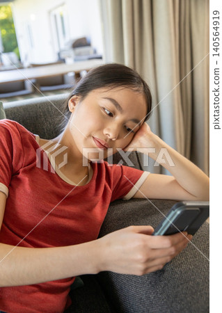 Asian woman reclining on sofa by window at home holding smartphone, decorative pillow Asian woman reclining on sofa by window at home holding smartphone, decorative pillow 140564919