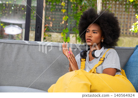 African american woman sitting on sofa by glass door in yellow overalls with headphones African american woman sitting on sofa by glass door in yellow overalls with headphones 140564924