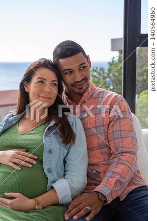 Diverse couple sitting on patio sofa by glass windows, man wrapping arm around pregnant belly Diverse couple sitting on patio sofa by glass windows, man wrapping arm around pregnant belly 140564960