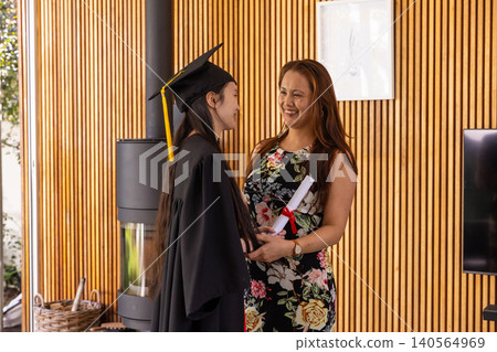 Asian mother-daughter standing in living room holding diploma wearing cap and gown Asian mother-daughter standing in living room holding diploma wearing cap and gown 140564969
