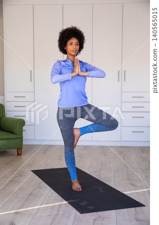 African american woman holding tree pose on black yoga mat in living room near armchair 140565015
