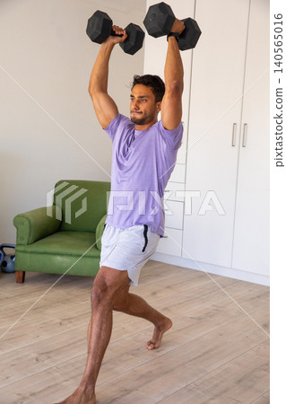 Man performing overhead lunge with hex dumbbells in home workout area next to kettlebell 140565016