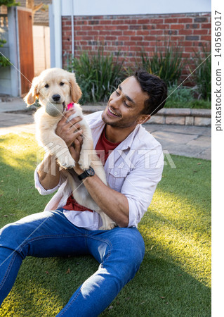 Man sitting in backyard lawn holding golden retriever puppy with collar with tennis ball Man sitting in backyard lawn holding golden retriever puppy with collar with tennis ball 140565017