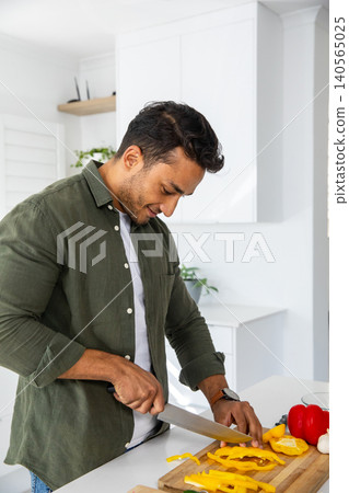 Man wielding knife slicing yellow bell pepper beside pepper on cutting board at kitchen counter Man wielding knife slicing yellow bell pepper beside pepper on cutting board at kitchen counter 140565025