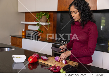 Indian woman slicing halved red onion on cutting board at kitchen island, copy space Indian woman slicing halved red onion on cutting board at kitchen island, copy space 140565030