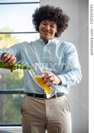 African american man pouring beer from green bottle into pint glass by large window African american man pouring beer from green bottle into pint glass by large window 140565045