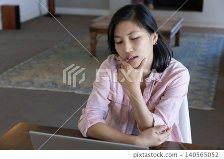 Asian woman in shirt working on laptop at desk at home with coffee table Asian woman in shirt working on laptop at desk at home with coffee table 140565052