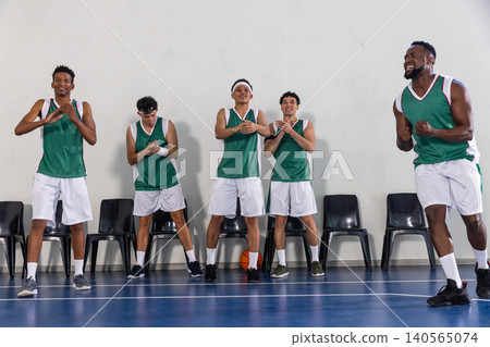 Diverse male basketball team in jerseys standing on court sideline clapping with ball near chairs Diverse male basketball team in jerseys standing on court sideline clapping with ball near chairs 140565074