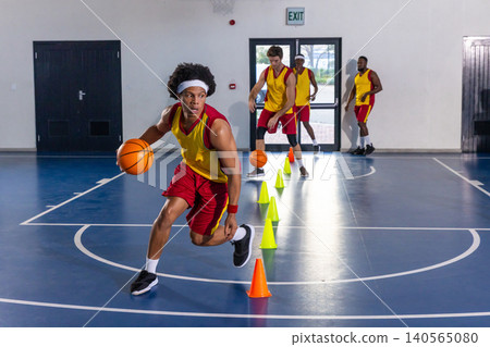 Diverse male basketball players dribbling balls around orange, green cones on court under hoop Diverse male basketball players dribbling balls around orange, green cones on court under hoop 140565080