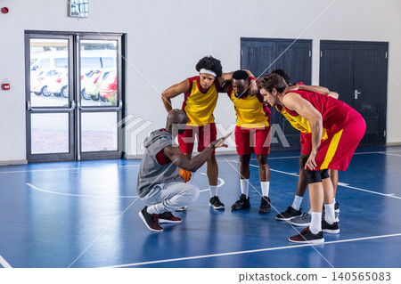 Diverse male basketball coach and team huddling on court holding orange ball directing play Diverse male basketball coach and team huddling on court holding orange ball directing play 140565083