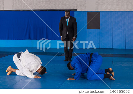Diverse martial artists bowing on blue mats at dojo in gis with referee in suit observing Diverse martial artists bowing on blue mats at dojo in gis with referee in suit observing 140565092